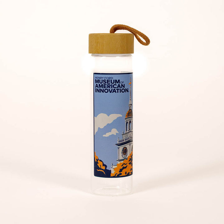 The Henry Ford Museum Building Glass Water Bottle features a clear design, bamboo lid, tan loop handle, and a label with a building illustration and "Henry Ford Museum of American Innovation" text.