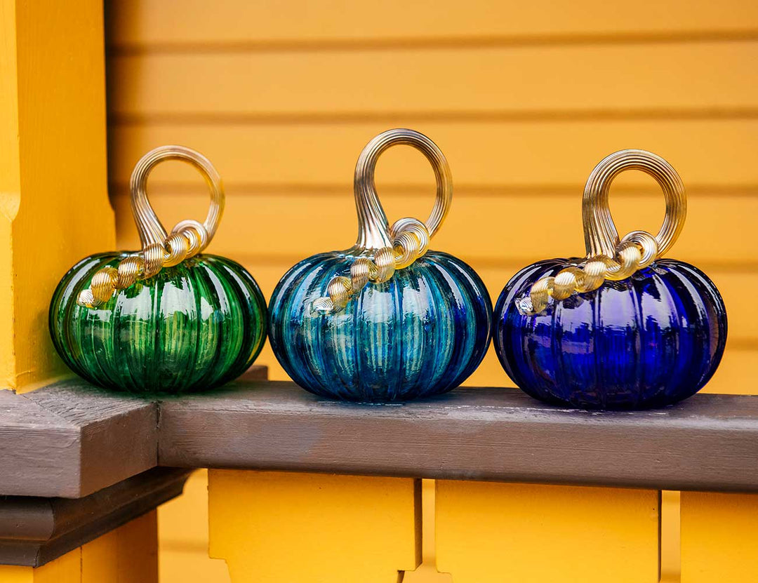 A Small Glass Pumpkin - Aqua with ribbed texture and a curly clear stem sits on a wooden ledge, evoking Greenfield Village charm against yellow siding and painted railings.