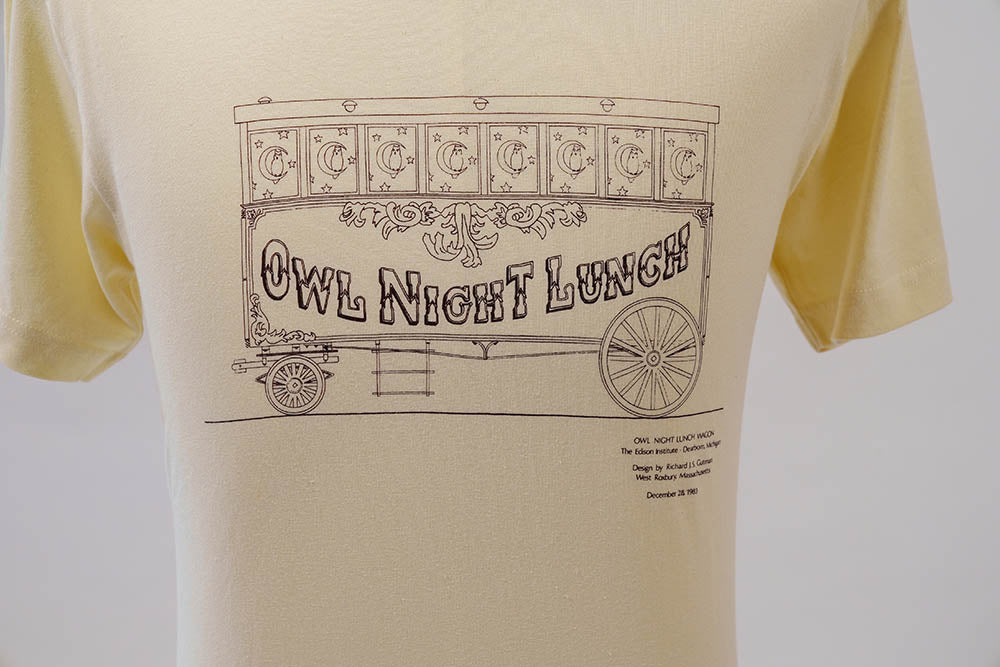 The Owl Night Lunch Wagon T-Shirt is yellow and features a vintage line drawing of the Owl Night Lunch Wagon with “Owl Night Lunch” in large decorative letters, inspired by Henry Ford’s Greenfield Village. Small text appears below the illustration on the right.