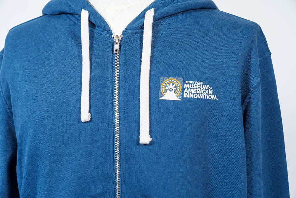 A close-up of The Henry Ford Museum Architecture Zip-Up Hooded Sweatshirt in blue, with white drawstrings and the museum’s logo on the left chest—an example of responsibly made apparel.
