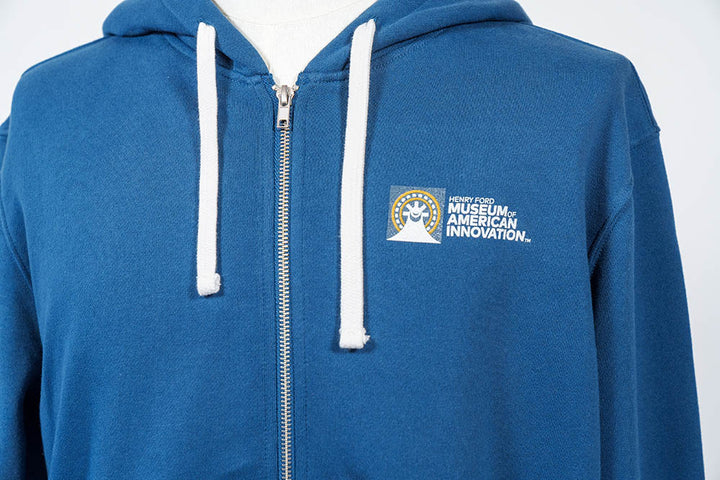A close-up of The Henry Ford Museum Architecture Zip-Up Hooded Sweatshirt in blue, with white drawstrings and the museum’s logo on the left chest—an example of responsibly made apparel.