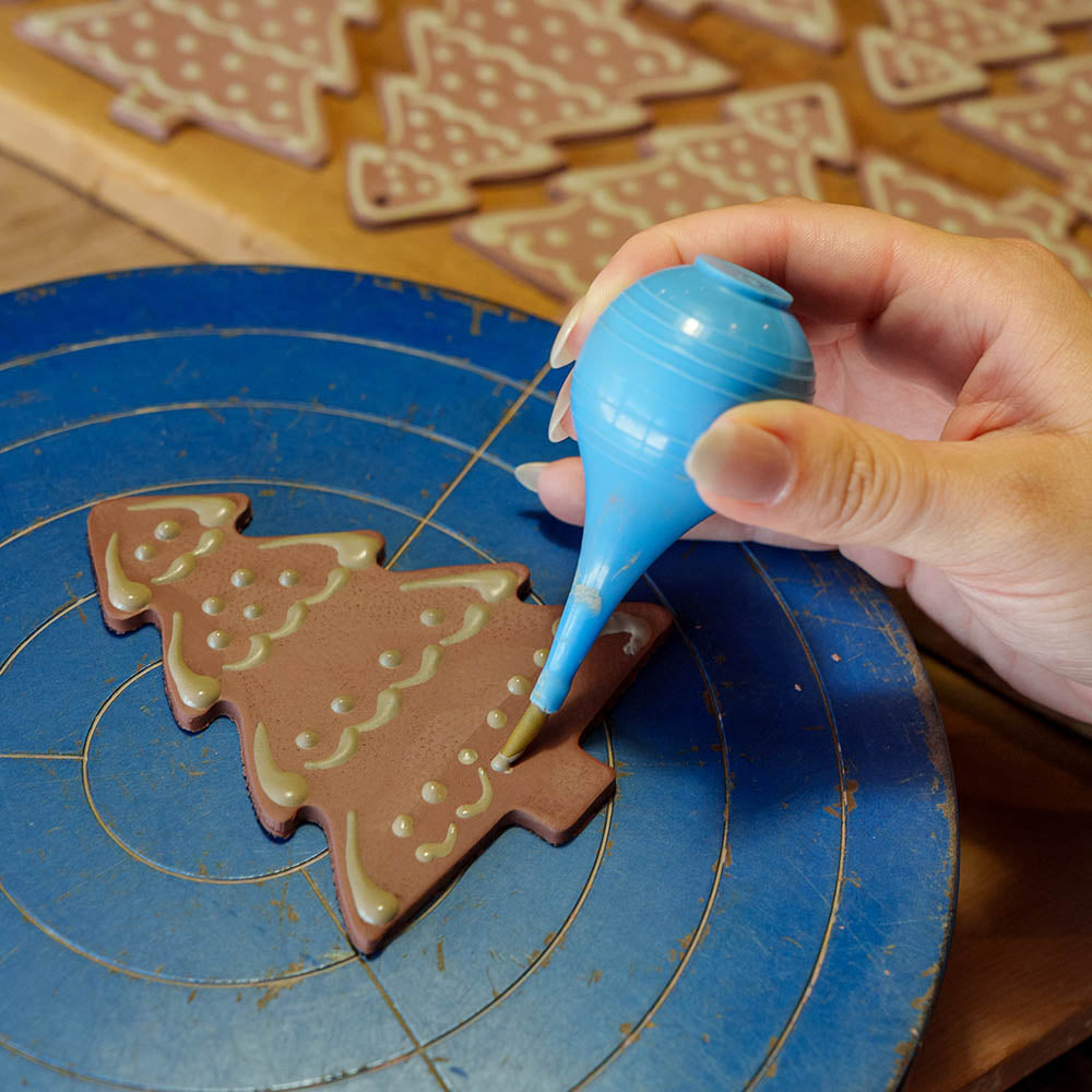 A hand decorates a chocolate Christmas tree cookie with blue icing, making it resemble the Redware Christmas Tree Ornament, while more undecorated cookies sit on a wooden table in the background.