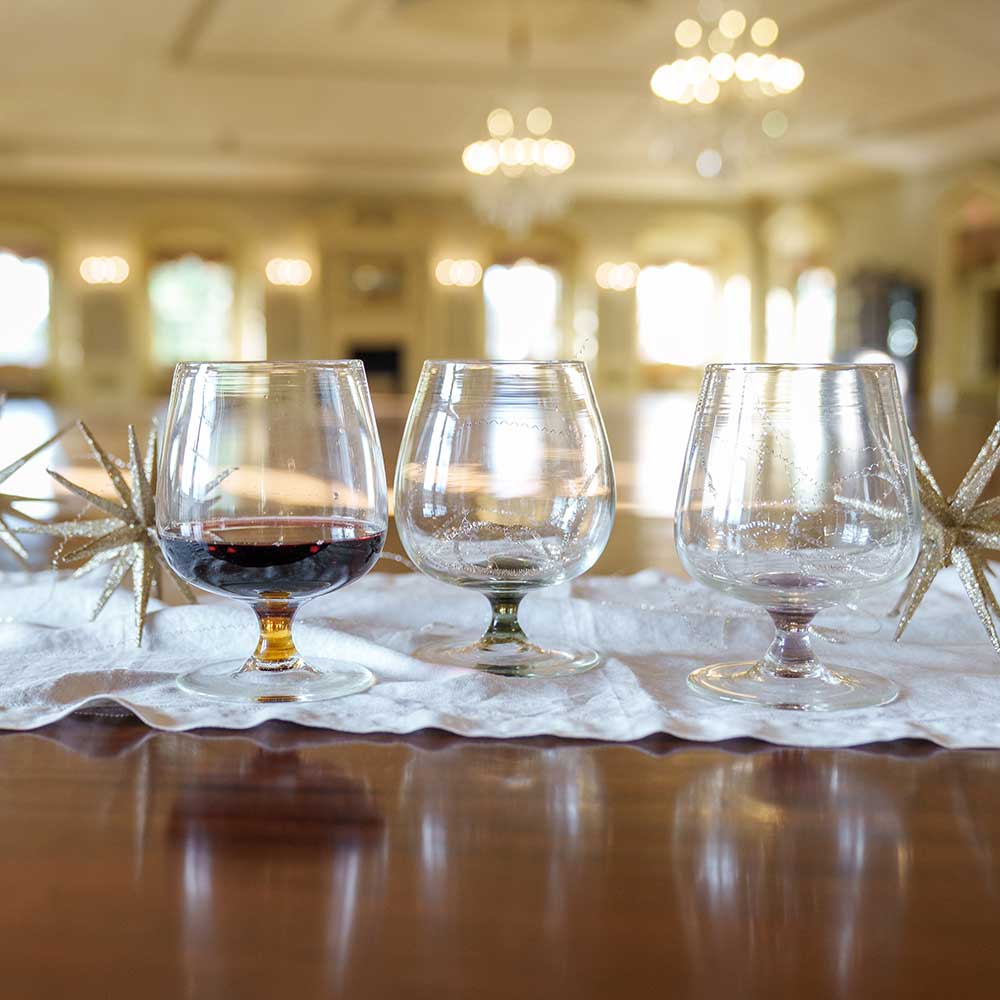 Three Mixology Short-Stemmed Red Wine Glasses, one with a hint of red wine, rest on a white cloth runner with gold star accents, adding elegance beneath sparkling chandeliers.