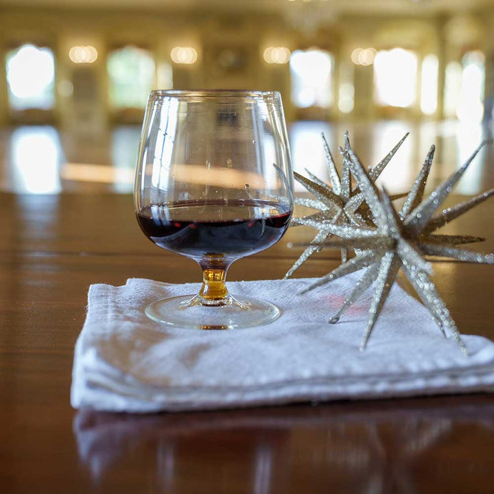 A Mixology Short-Stemmed Red Wine Glass rests on a folded white napkin atop a wooden table beside two decorative silver star ornaments, with a softly blurred background bathed in warm natural light.