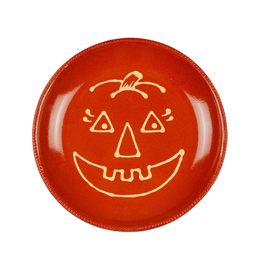The Pumpkin Plate is a round orange redware plate featuring a yellow-outlined pumpkin face with triangle eyes, nose, and a wide toothy smile. Hand decorated with a smooth, glossy finish.