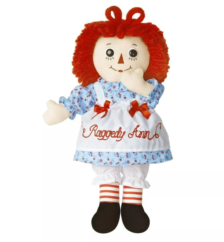 Raggedy Ann doll featuring red yarn hair, a white apron labeled “Raggedy Ann,” blue floral dress, red-striped socks, black shoes, button eyes, and a stitched smile.