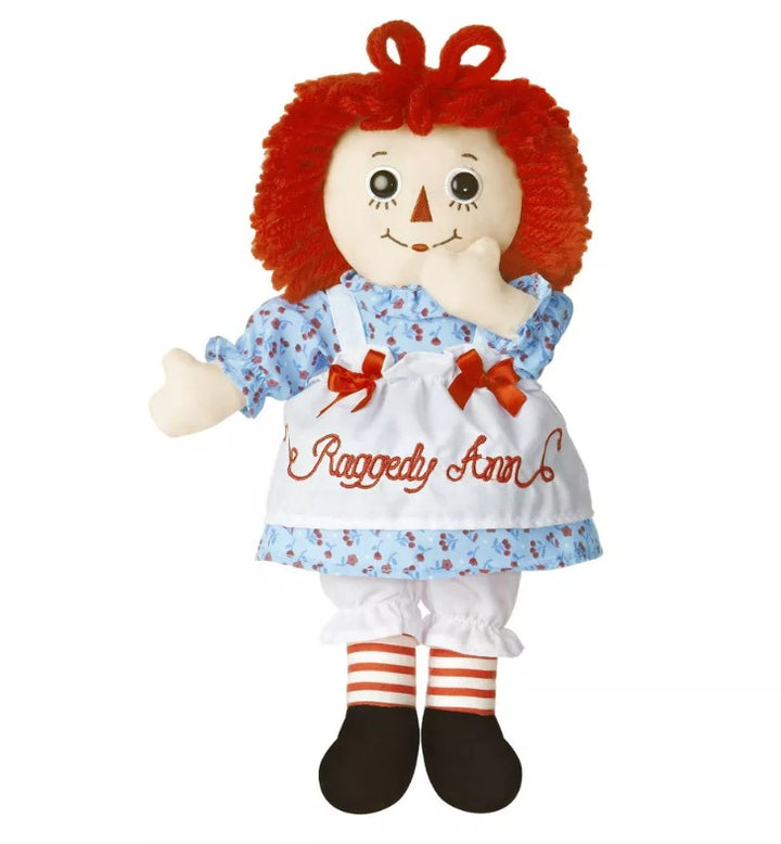Raggedy Ann doll featuring red yarn hair, a white apron labeled “Raggedy Ann,” blue floral dress, red-striped socks, black shoes, button eyes, and a stitched smile.