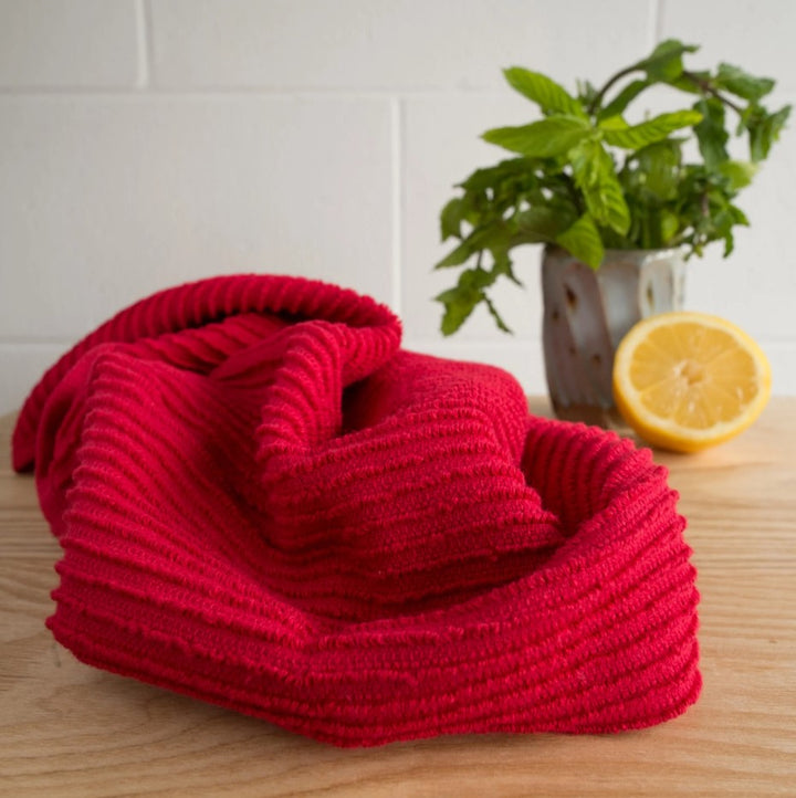 The Ripple Red Dishtowel - Single, crafted in Turkey, rests on a wooden surface near a lemon half and fresh mint in a vase, set against a white tiled wall.
