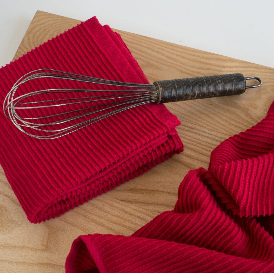 A Ripple Red Dishtowel - Single, made in Turkey, lies folded on a light wooden surface with a metal whisk atop it; another matching, highly absorbent dishtowel appears partially in the foreground.