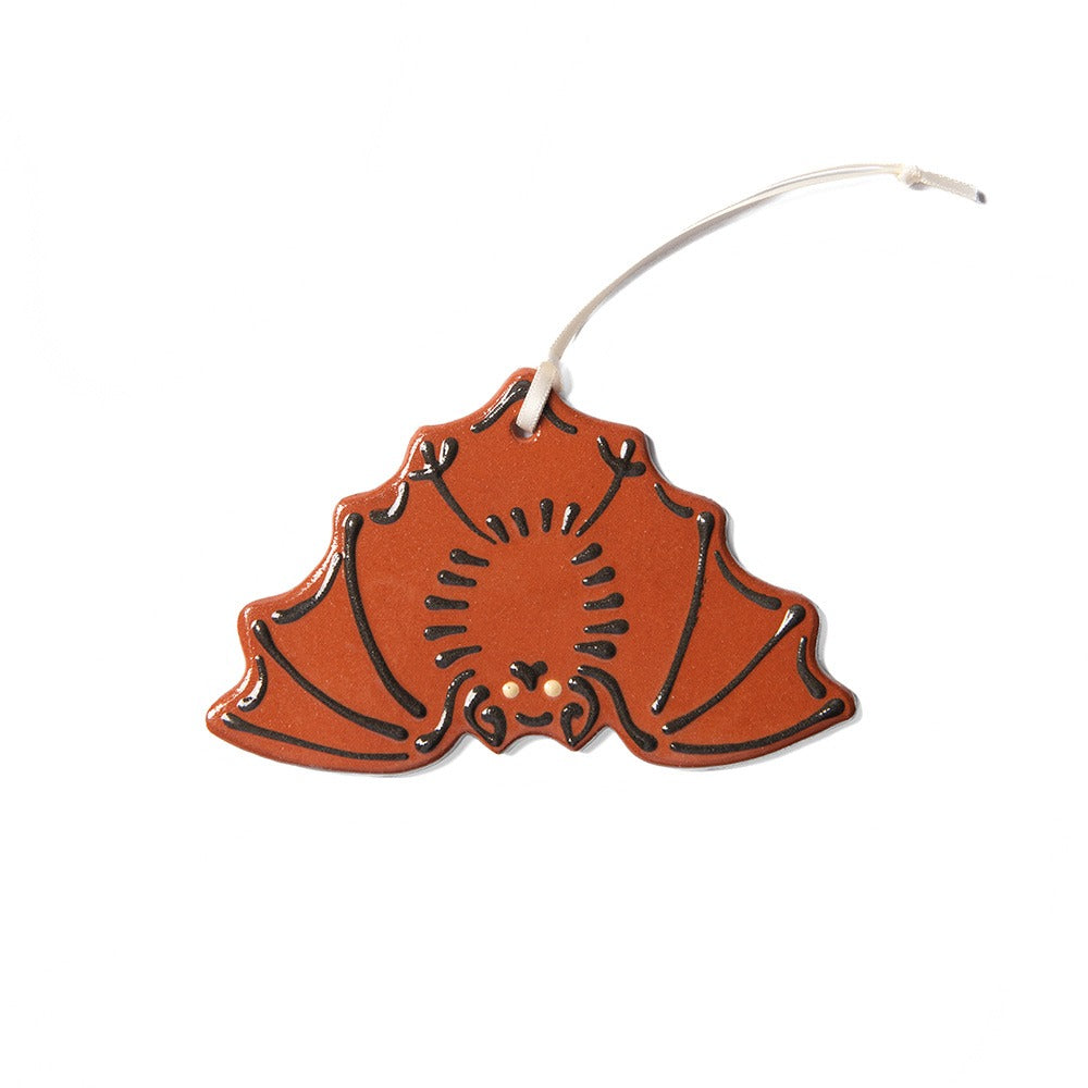The Redware Bat Ornament is an orange, bat-shaped decoration made from hand-crafted redware clay, featuring black outlines and a white hanging string, shown against a white background.