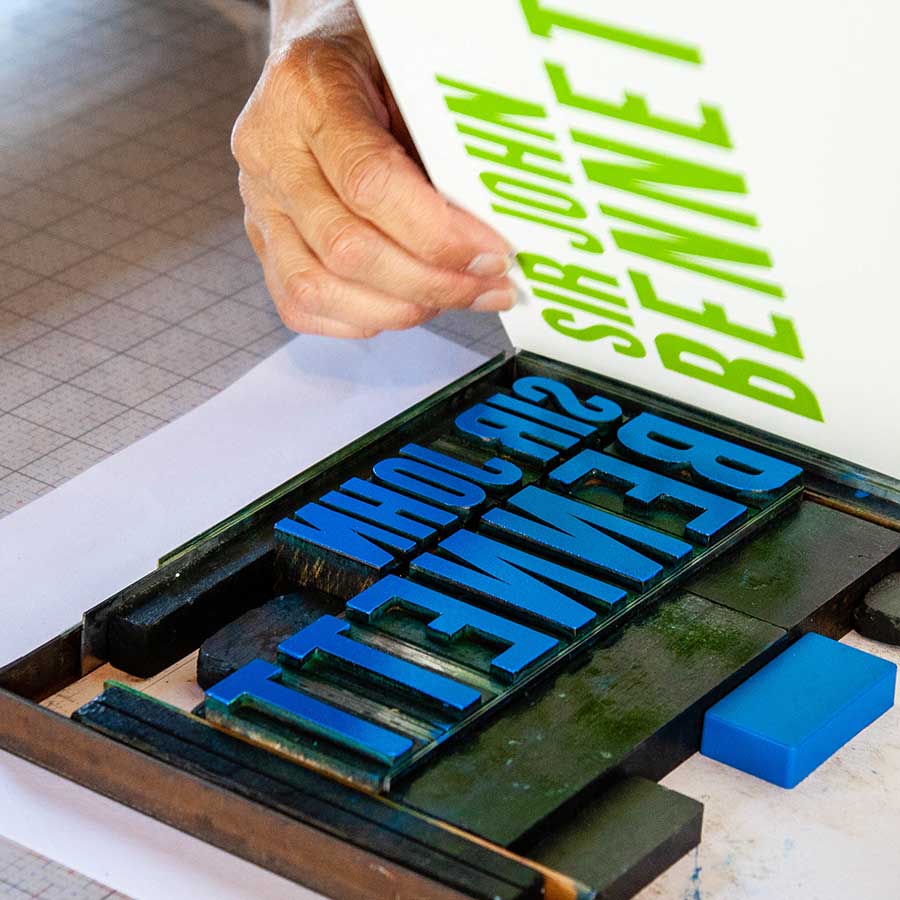 A person lifts the Sir John Bennett 8.5"x11" print—handcrafted on a letterpress with blue raised letters and green ink, using historic wood type—revealing a grid-patterned design.