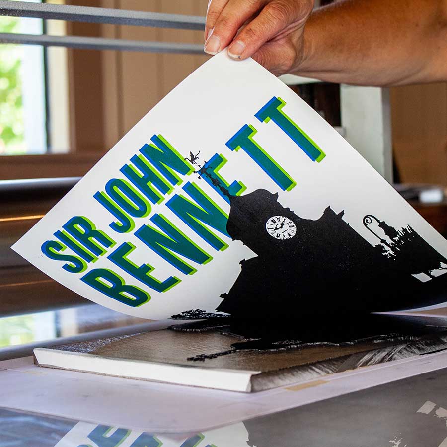 A hand holds the Sir John Bennett 8.5"x11" print, a vibrant limited-edition artwork featuring "SIR JOHN BENNETT" above a clock tower silhouette, created with historic wood type as sunlight streams through a window behind.