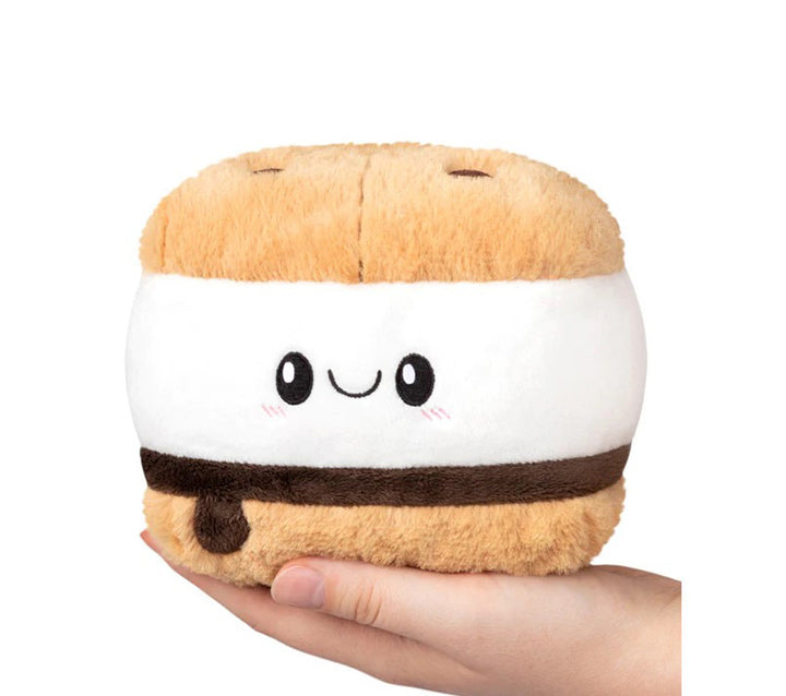 A hand holds the Plush Squishable S'more Snacker, a cute plush toy shaped like a s'more with tan, white, and brown layers plus a smiling face on the white middle—perfect for snack lovers who want bite-sized plush fun.