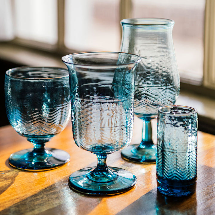 Four Steel Blue American Heritage Firing Glass cups, each uniquely shaped and textured, are arranged on a wooden table with sunlight streaming through a nearby window.