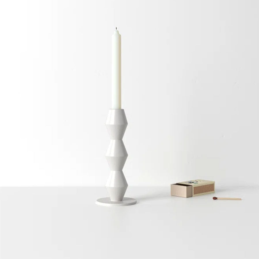 The Vector Tabletop Metal Candlestick with a tall white taper candle stands on a white surface beside an open matchbox and a single matchstick, set against a minimalistic, plain background.