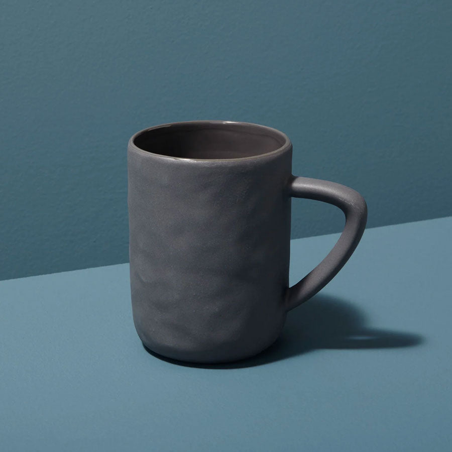 The Tam Stoneware Mug - Slate, handcrafted by women artisans in Vietnam, features a matte gray finish and large handle. It sits on a blue surface, casting a soft shadow. Dishwasher and microwave safe.