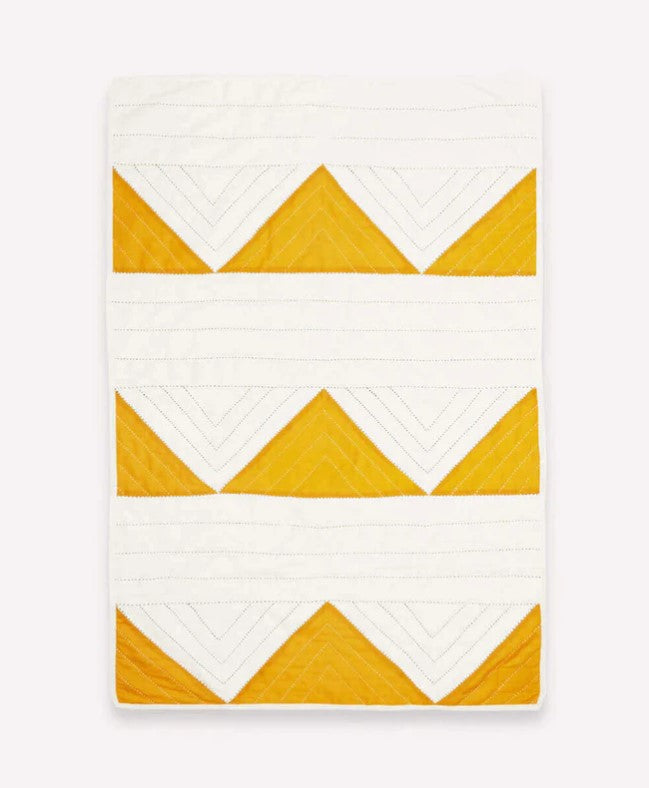 The Mustard Triangle Throw Blanket is hand quilted with a geometric chevron and triangle pattern in white and mustard yellow—outlined with stitched lines, making it perfect as embroidered nursery decor.
