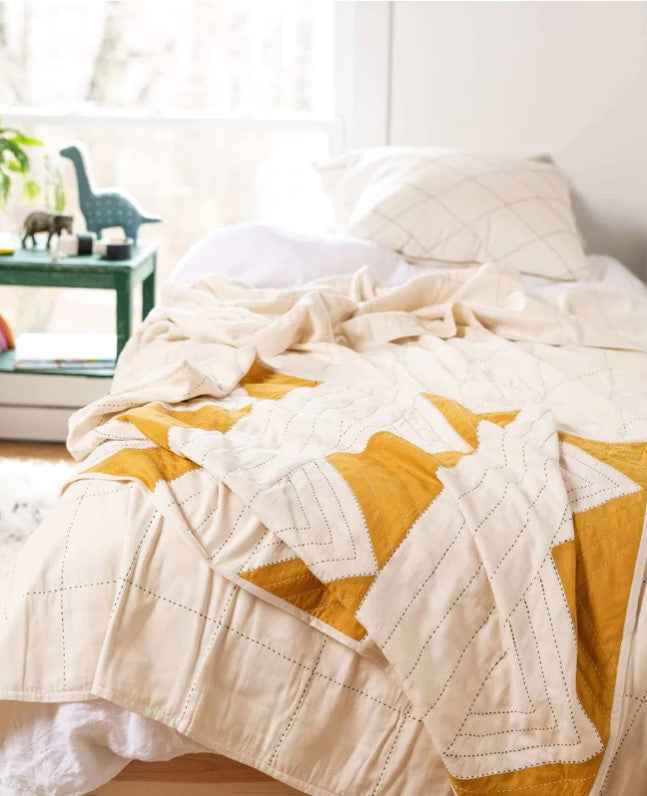 A cozy, unmade bed features the Mustard Triangle Throw Blanket in cream and yellow with matching pillows by a sunny window. A green side table with decor, including a dinosaur figure, adds playful charm beside the bed.