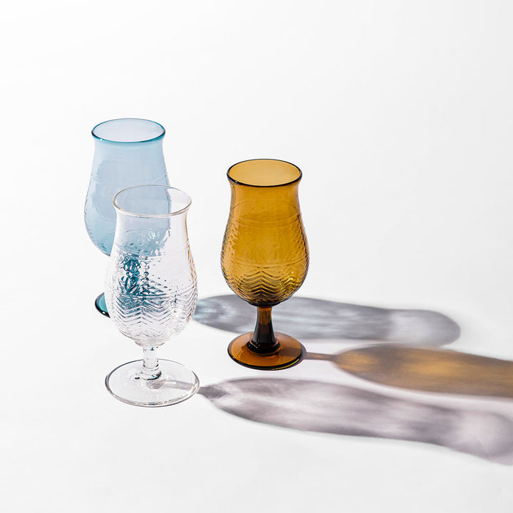 The American Heritage Molded Tulip Glass in Steel Blue is displayed alongside clear and amber goblets on a white surface, casting colorful shadows and bringing elegance to any table setting.