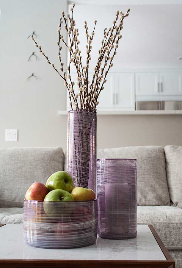 The Matassa Tall Cylinder Vase (Multiple Colors) with tall pussy willow branches sits on a marble table beside a bowl of apples and another glass vase, in front of a beige sofa and a white kitchen backdrop.