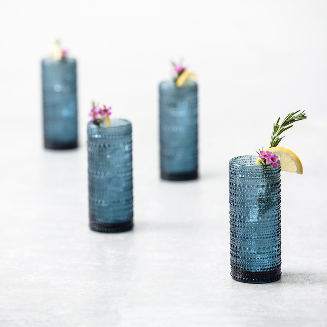 Four Jupiter Collins Highball 10.8oz Glasses in Cornflower, each filled with a refreshing drink and garnished with lemon, rosemary, and pink flowers, are arranged in a staggered line on a light surface.