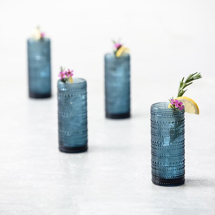 Four Jupiter Collins Highball 10.8oz Glasses in Cornflower, each filled with a refreshing drink and garnished with lemon, rosemary, and pink flowers, are arranged in a staggered line on a light surface.