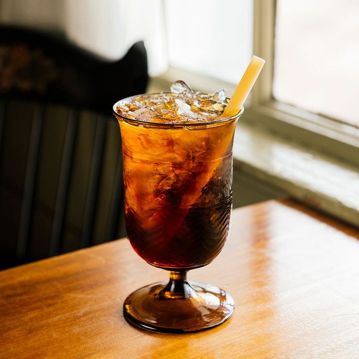 Sunlight illuminates a glass of iced tea with a straw, served in the American Heritage Molded Water Goblet - Tea, on a wooden table near a window.