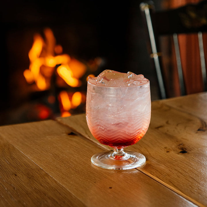 A glass of iced pink and red drink sits on a wooden table in an American Heritage Molded Wine Goblet - Clear, with a blurred fireplace and orange flames in the background, creating a warm, cozy atmosphere.