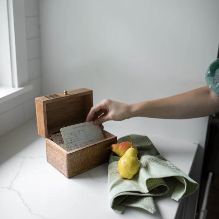 A hand opens the Carved Wood Recipe Box on a white countertop beside a green cloth and two fruits, illuminated by soft daylight from a nearby window.