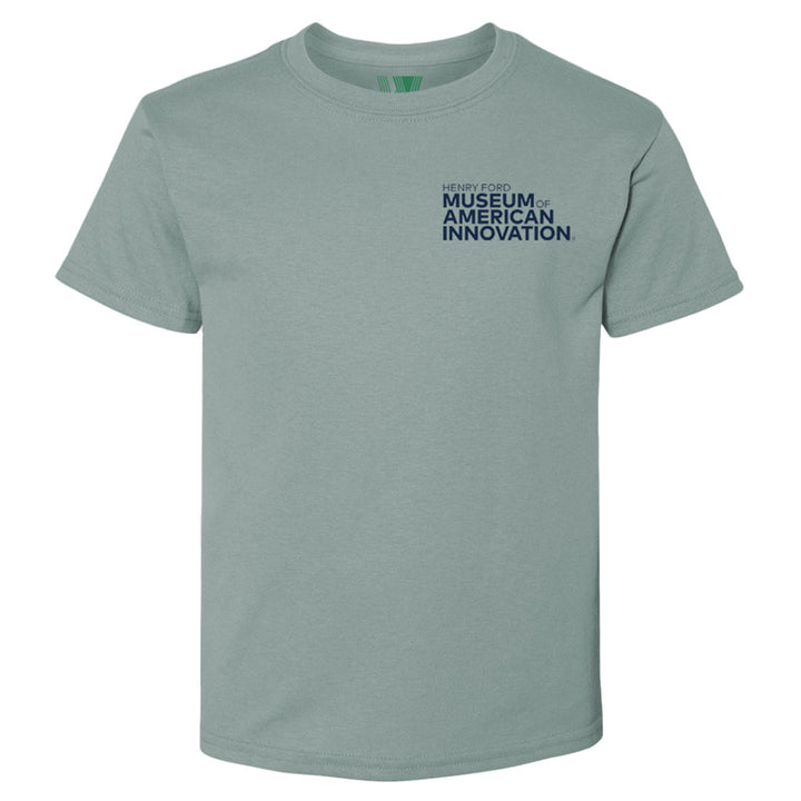 The Henry Ford Museum Clocktower T-Shirt in light gray-green organic cotton features “HENRY FORD MUSEUM OF AMERICAN INNOVATION” in dark blue on the left chest. Shown on a plain white background.