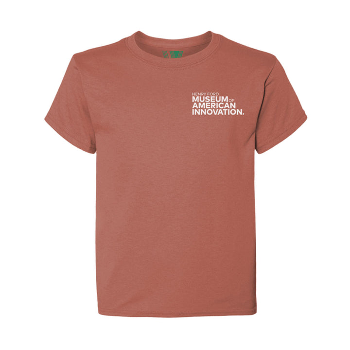 The Henry Ford Museum Building Clocktower Youth T-Shirt is a rust-colored organic cotton tee with white text on the upper left chest, displayed on a plain white background.