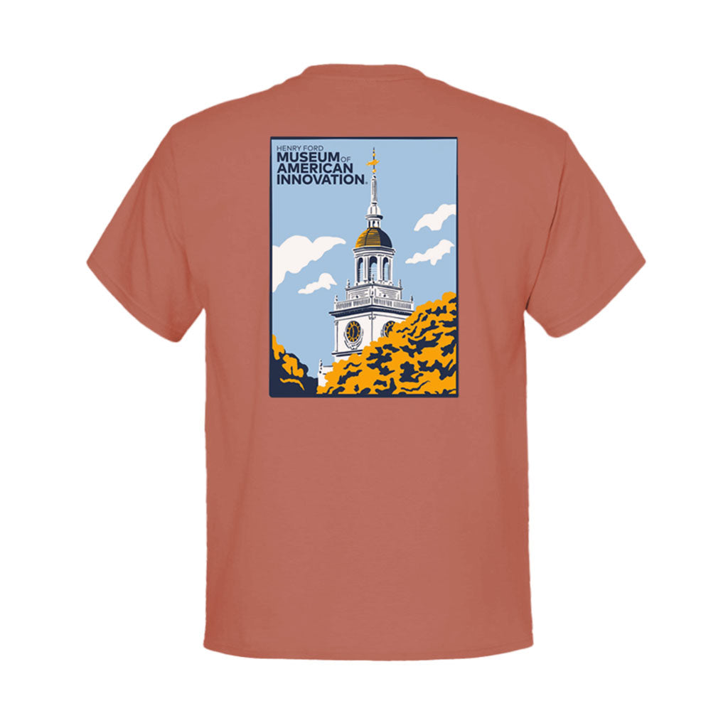 The Henry Ford Museum Building Clocktower Youth T-Shirt is rust-colored with custom artwork of a white clock tower with a gold spire over green and yellow foliage, and "Museum of American Innovation" in the top left corner.