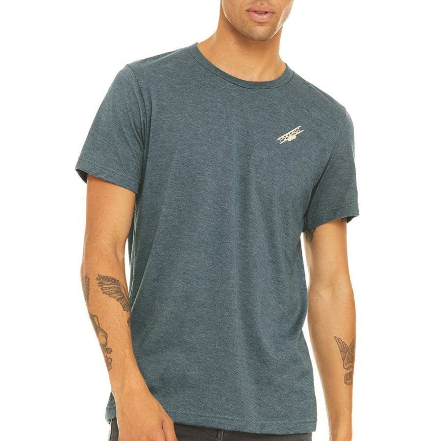 A person with tattooed arms wears a fitted, short-sleeve, heathered dark blue Greenfield Village Model Wright Brothers Biplane T-Shirt featuring a small white logo on the left chest, standing against a plain background.