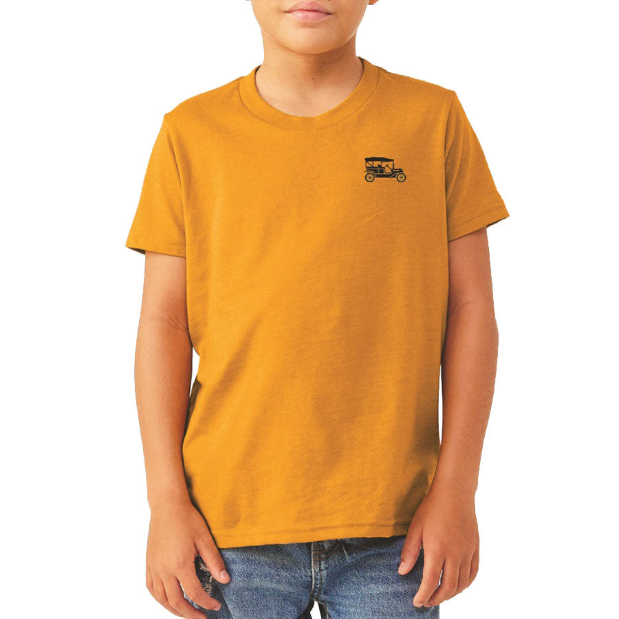 A child wears the Youth Greenfield Village Model T Adventures T-Shirt in mustard yellow, featuring a small black vintage car graphic on the left chest, paired with blue jeans—ideal for young Greenfield Village enthusiasts.
