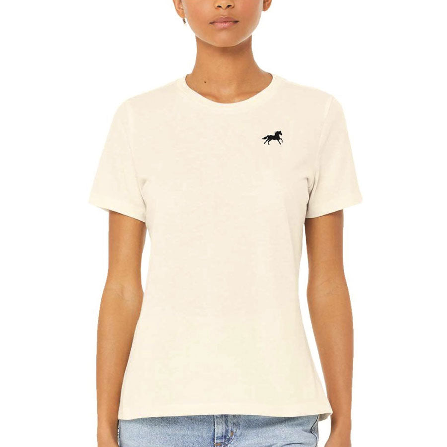 A person wearing the Ladies Greenfield Village Wild at Heart T-Shirt, featuring a cream color with horse embroidery and a small black horse logo on the left chest, paired with light blue jeans against a plain background.